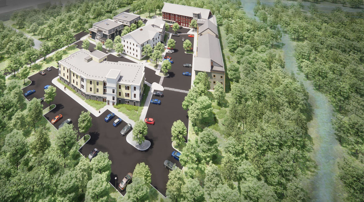 Aerial view shows buildings with parking surrounded by woods with a small river behind.